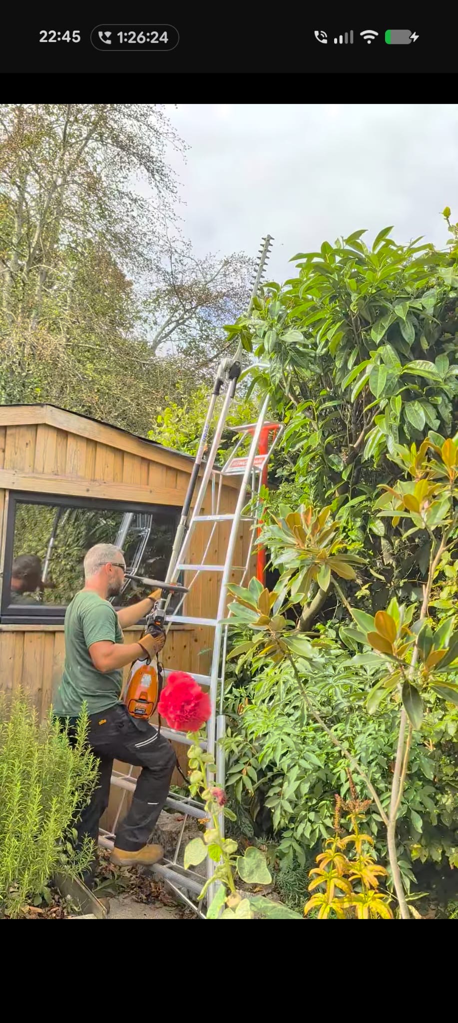 Mitch using a long-reach hedge trimmer beside a garden building